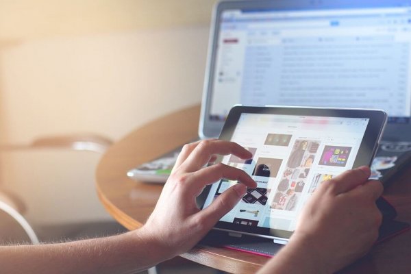 A person using an iPad with a laptop in the background